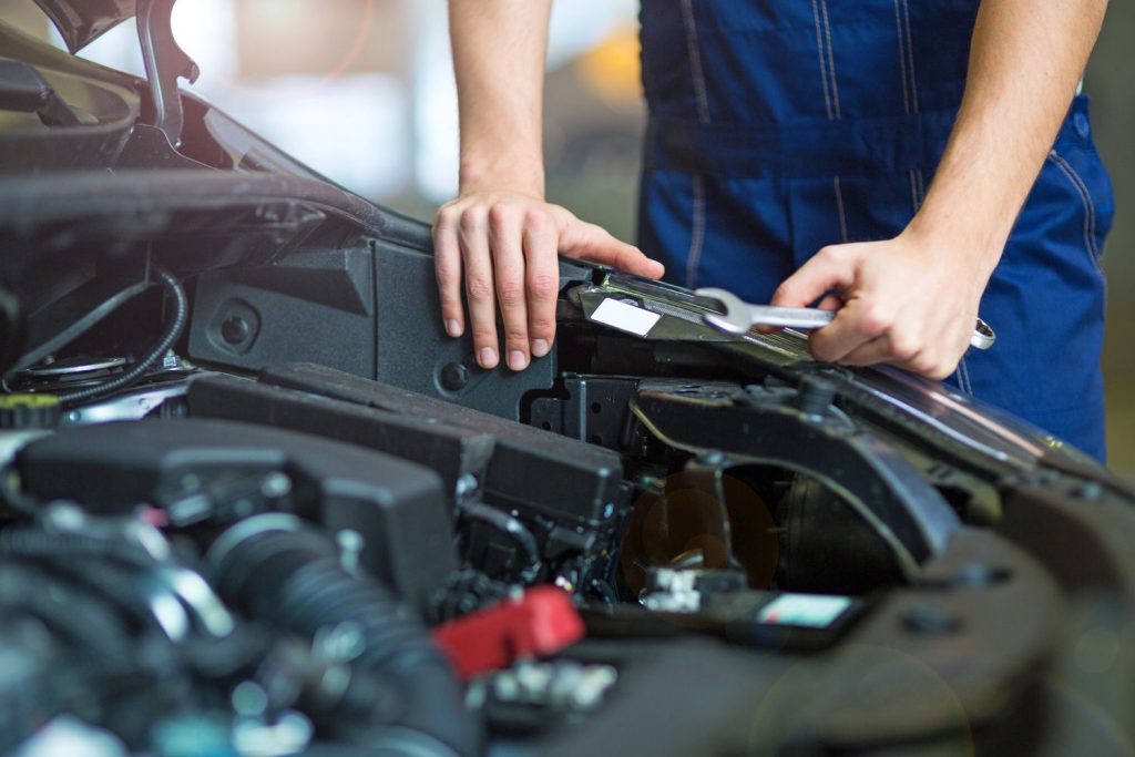 Mechanic performing third party inspection in open car engine bay, using wrench to adjust components.