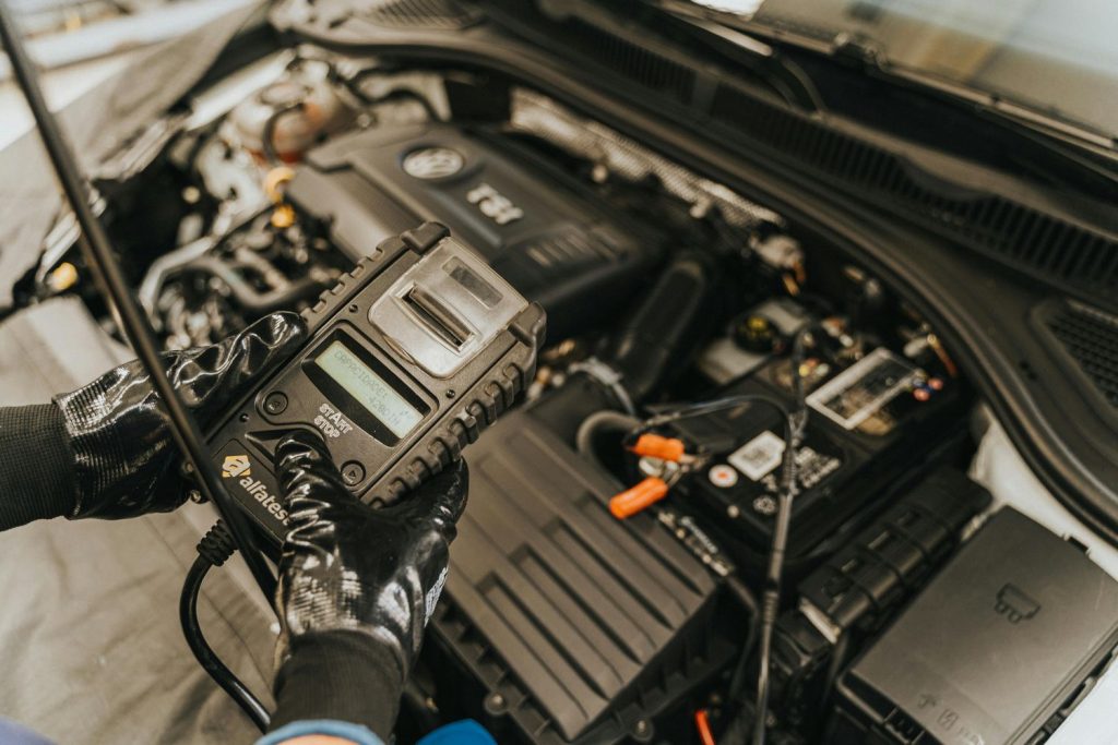 Technician conducting third party inspection on car battery using handheld diagnostic tool in engine bay.