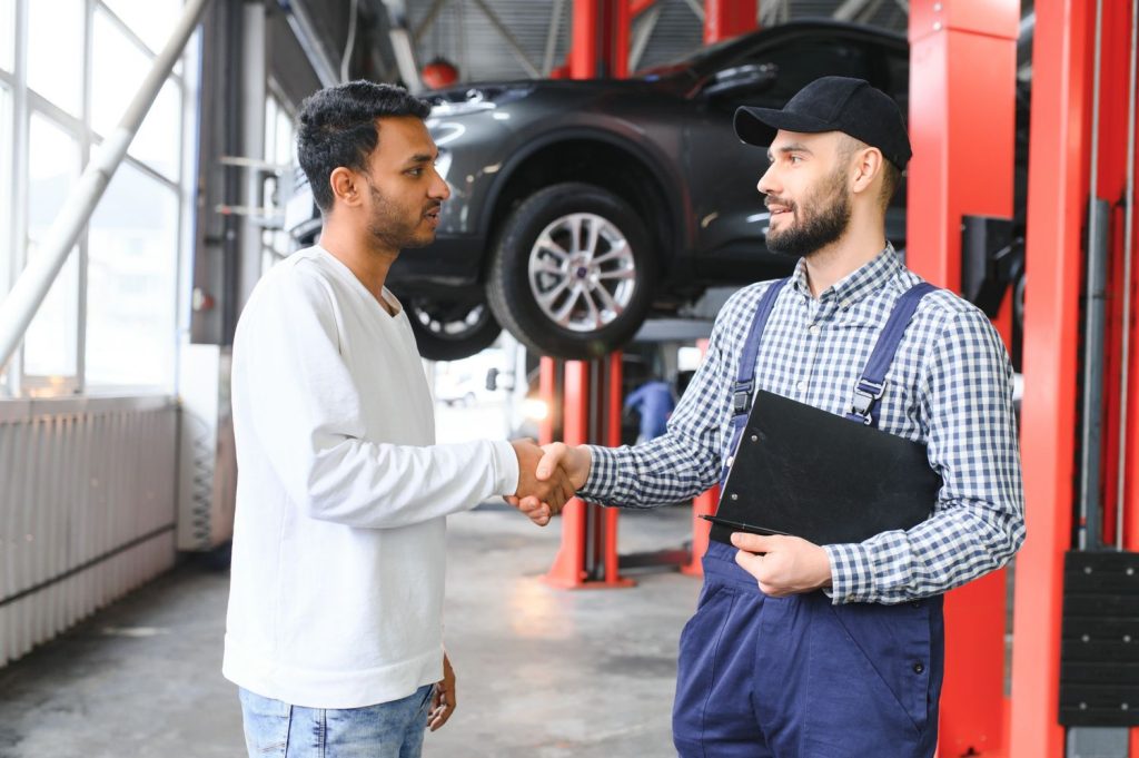 A client shakes hands with a mechanic from the best car inspection service Houston, with a lifted car in the background.