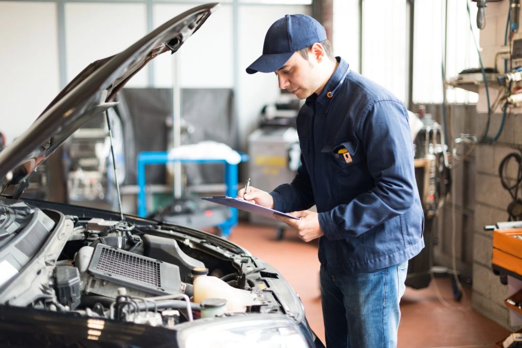 A mechanic conducts a pre purchase used car inspection Houston, taking notes on a clipboard beside an open car engine in a garage.