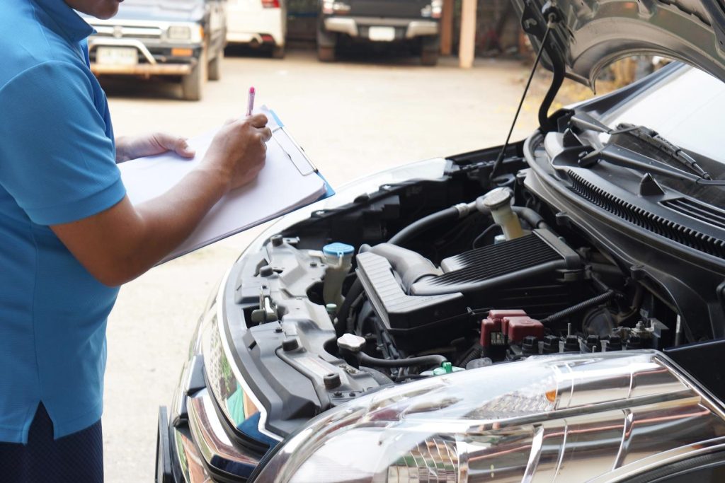 A mechanic in blue shirt conducting a third party inspection Houston on a car engine with a clipboard.