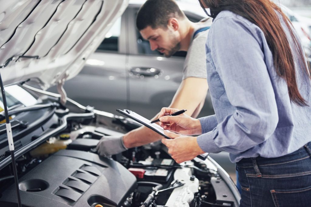 Professional third party inspection of a car's engine by mechanic and inspector with clipboard in garage.