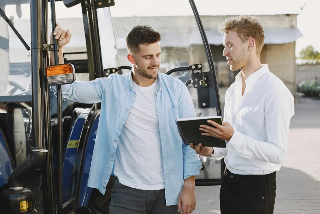 Two men discussing pre-purchase used car inspection in Houston near a tractor, one holding a tablet.