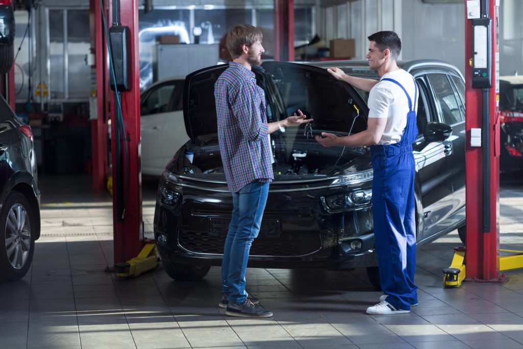 A mechanic explains engine details to a client at the best car inspection service Houston, with an open car hood in a garage.