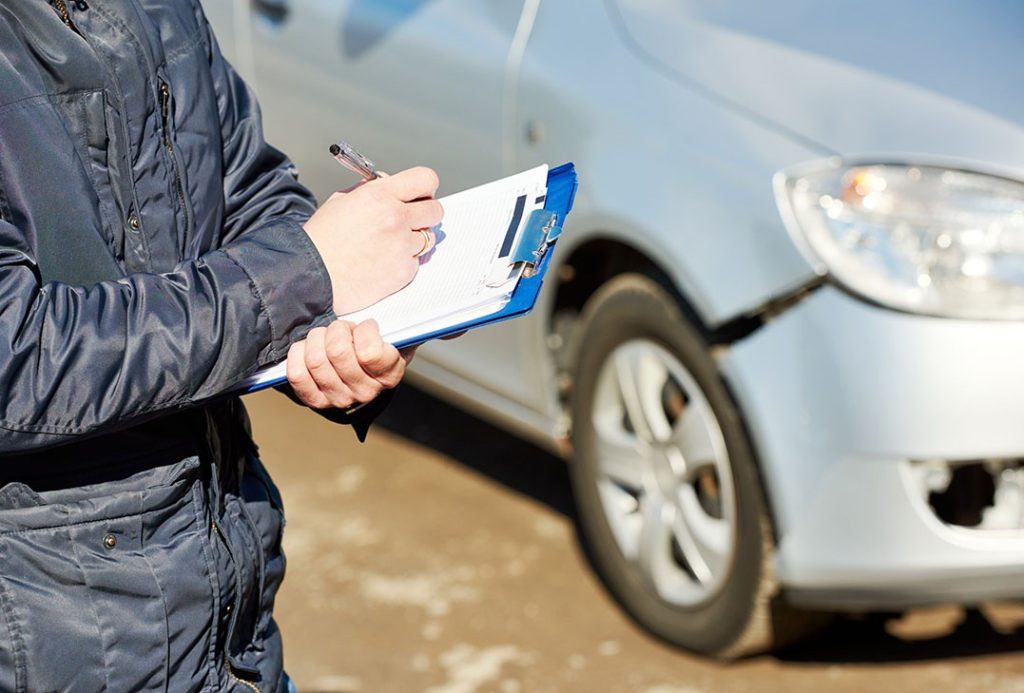Professional certified third party car inspector writing notes on a clipboard near a damaged silver car.