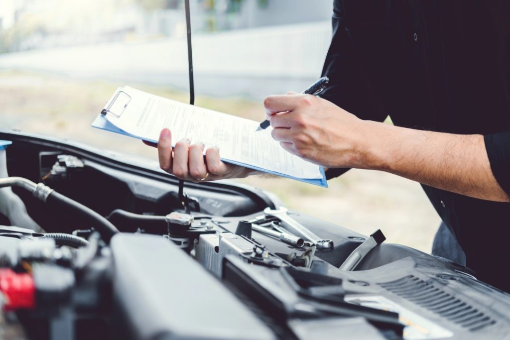 Mechanic using a checklist for an independent used car inspection while examining the engine bay of a sedan.