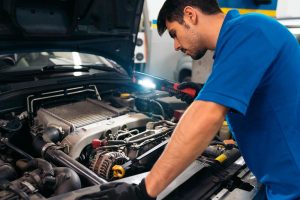 A certified mobile mechanic uses a bright work light to inspect intricate engine details under the open hood of a used car.