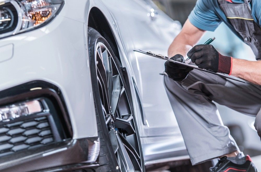 An inspector in workwear takes notes on a clipboard during a professional Pre purchase vehicle inspection Houston of a white car’s wheel.