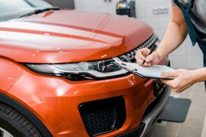 A mechanic takes notes in front of an orange-red SUV during a detailed Pre purchase vehicle inspection Houston.