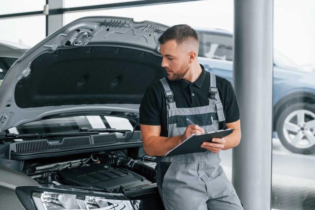 Mechanic filling out a third party inspection checklist next to an open car hood at an auto dealership.