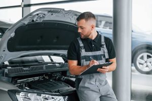 Mechanic filling out a third party inspection checklist next to an open car hood at an auto dealership.