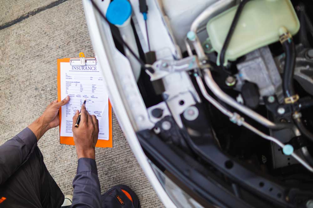Mechanic filling out paperwork during a certified pre owned car inspection service on an open vehicle engine.